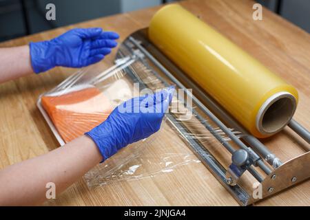 Un employé portant des gants jetables bleus enveloppe un morceau de filet de saumon allongé sur un plateau en plastique blanc dans un film transparent. Concept de stockage, de conservation salée et Banque D'Images