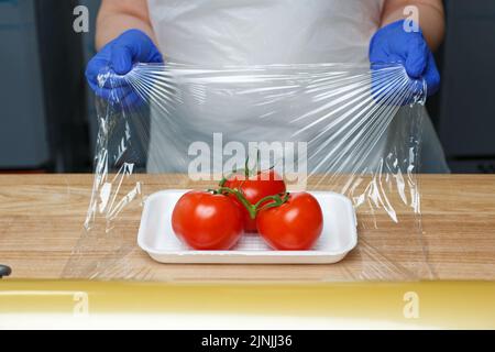 Détaillants d'épicerie. Emballage des produits alimentaires. Un employé inconnu en gants jetables bleus encapsule dans un film transparent alimentaire tomates allongé sur du plastique blanc t Banque D'Images