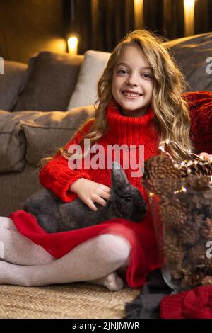 Une petite fille blonde avec un lapin gris dans ses bras à côté de la cheminée. Le concept des vacances d'hiver est Noël et jour de l'an. Fête magique Banque D'Images