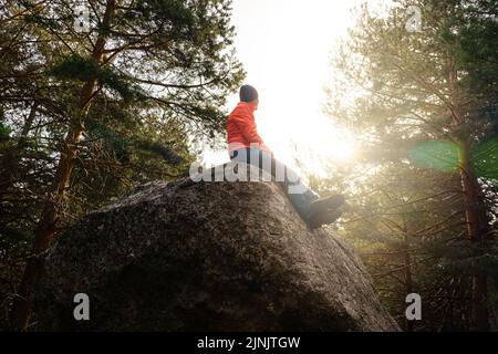 Homme perché sur un grand rocher dans la forêt, regardant le soleil au lever du soleil avec une lumière dorée et une atmosphère calme. Banque D'Images