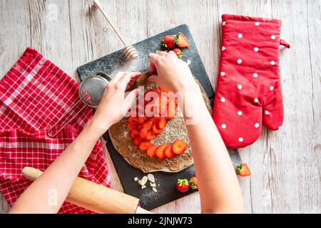 Galette avec préparation de fraises, détail des mains de cuisinière. Gâteau à tarte placé sur un bureau noir et une table en bois avec des outils de cuisson sur le côté. Banque D'Images