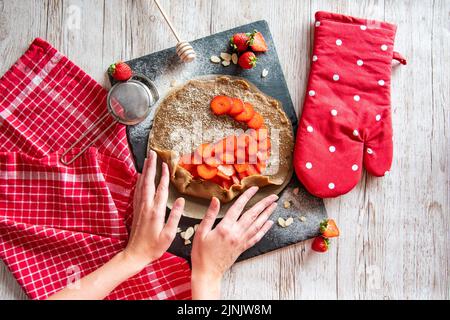 Galette avec préparation de fraises, détail des mains de cuisinière. Gâteau à tarte placé sur un bureau noir et une table en bois avec des outils de cuisson sur le côté. Banque D'Images