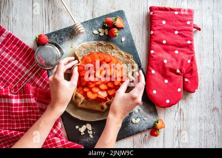 Galette avec préparation de fraises, détail des mains de cuisinière. Gâteau à tarte placé sur un bureau noir et une table en bois avec des outils de cuisson sur le côté. Banque D'Images