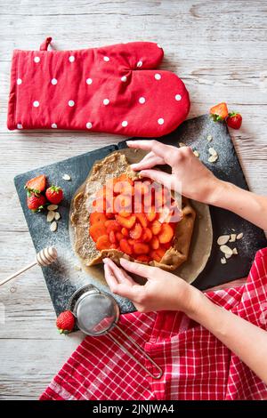 Galette avec préparation de fraises, détail des mains de cuisinière. Gâteau à tarte placé sur un bureau noir et une table en bois avec des outils de cuisson sur le côté. Banque D'Images