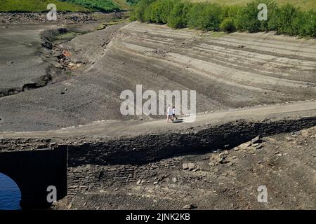 Les gens marchent à travers la terre sèche fissurée au réservoir Baitings à Ripponden, West Yorkshire, où les niveaux d'eau sont considérablement faibles. Le stock total d'eau dans les réservoirs d'Angleterre à la fin du mois de juillet était de 65 pour cent de sa capacité normale - le niveau le plus bas pour ce point de l'année civile depuis 1995, a déclaré l'Agence de l'environnement. Date de la photo: Vendredi 12 août 2022. Banque D'Images