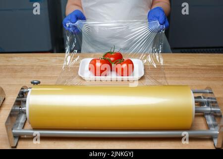 Détaillants d'épicerie. Emballage des produits alimentaires. Un employé inconnu en gants jetables bleus encapsule dans un film transparent alimentaire tomates allongé sur du plastique blanc t Banque D'Images