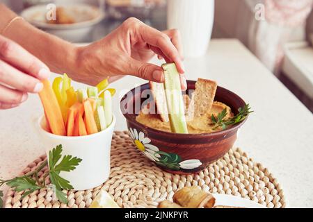 Les mains de la femme trempent le concombre frais dans du houmous maison. Biscuits, persil et légumes frais sur la table. En-cas sains végétariens rustiques en gros plan Banque D'Images