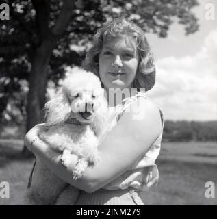 1960s, historique, une jeune femme debout à l'extérieur tenant son chien d'animal de compagnie, un petit caniche, Angleterre, Royaume-Uni, connu comme un jouet caniche. Race de chiens aquatiques, les poddles sont des chiens intelligents, alertes et actifs. Banque D'Images