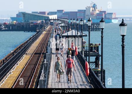 Southend on Sea, Essex, Royaume-Uni. 12th août 2022. Le temps chaud s'est poursuivi dans la nouvelle ville côtière de Southend sur la mer, avec de nombreuses personnes se dirigeant vers la station pour se rafraîchir par la mer. Les gens marchent sur la longueur de Southend Pier tandis qu'un énorme containership passe la fin Banque D'Images