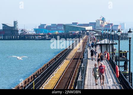 Southend on Sea, Essex, Royaume-Uni. 12th août 2022. Le temps chaud s'est poursuivi dans la nouvelle ville côtière de Southend sur la mer, avec de nombreuses personnes se dirigeant vers la station pour se rafraîchir par la mer. Les gens marchent sur la longueur de Southend Pier tandis qu'un énorme containership passe la fin Banque D'Images