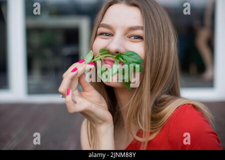 Portrait d'une jeune fille souriante et mignonne avec des cheveux longs et équitables portant un T-shirt rouge, tenant un basilic vert frais comme une moustache. Banque D'Images