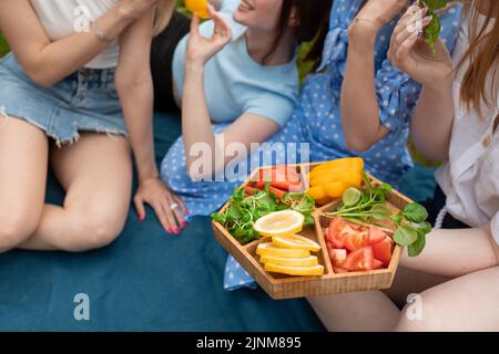 Photo rognée de jeunes femmes mangeant, tenant des légumes coupés, assis sur un écossais bleu sur le pique-nique. Amitié, alimentation saine. Banque D'Images