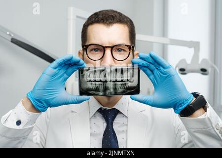 Un dentiste masculin recouvre sa bouche avec un smartphone avec des rayons X de ses dents sur un écran d'appareil. Banque D'Images