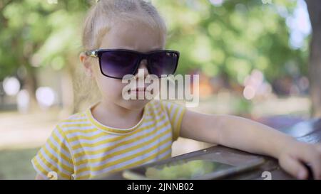 Petite fille dans les lunettes de soleil de mamans apprend la langue étrangère en répétant des mots à partir du téléphone portable. Portrait en gros plan d'une fille assise en ville Banque D'Images