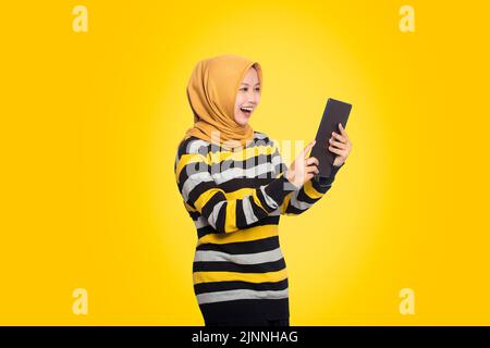 Belle jeune femme musulmane stupéfait de tablette. Bonne adolescente en robe rayée utilisant un ordinateur de tablette, isolé sur fond jaune. Banque D'Images