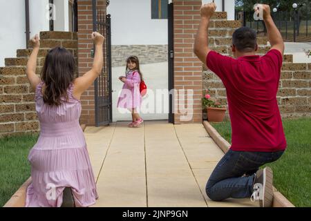 Image d'un couple de parents qui applaudissent pour leur fille avec un sac d'école et un tablier rose allant au premier jour de l'école. Banque D'Images