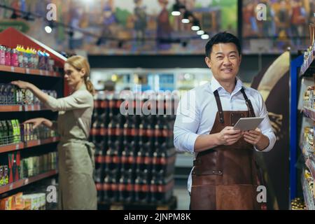 Portrait d'un vendeur asiatique réussi et heureux dans un supermarché, un homme dans un tablier regarde la caméra et souriant, le directeur de magasin tient une tablette pour l'examen des produits Banque D'Images