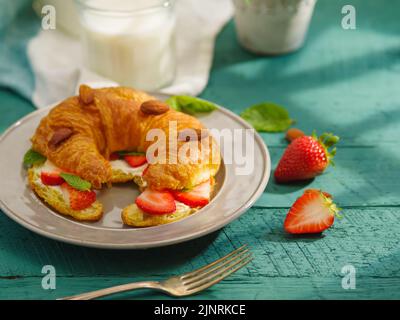 Croissant français frais avec fraises mûres, amandes et feuilles de menthe parfumées, lait dans un pot et une tasse de café sur fond turquoise. Français b Banque D'Images