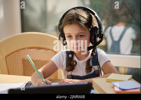 Belle jeune fille écolière portant un casque audio sans fil pour regarder une vidéo éducative tout en étudiant à distance depuis la maison Banque D'Images