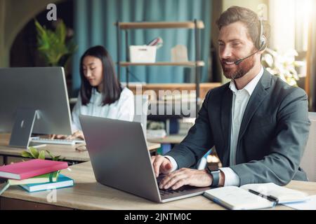 Opérateur masculin utilisant un casque et un ordinateur tout en discutant avec des clients en ligne sur l'arrière-plan des collègues Banque D'Images