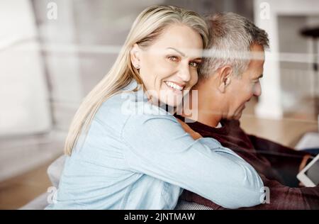 Quand vous trouvez votre roi se tenir sur lui. Une femme mûre embrassant son mari pendant qu'il utilise une tablette numérique sur le canapé à la maison. Banque D'Images