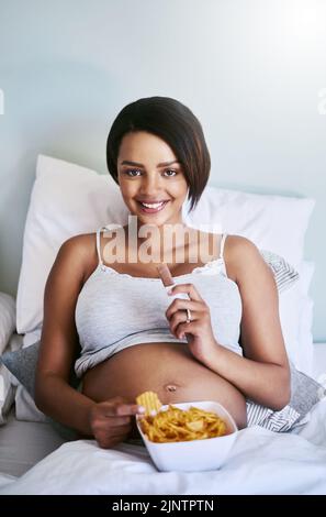 Le bébé m'a fait manger. Une femme enceinte appréciant un chocolat et des chips de pomme de terre au lit. Banque D'Images