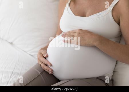 La femme enceinte met les paumes sur le ventre assis sur le lit, en gros plan Banque D'Images