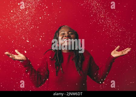 Femme joyeuse avec des dreadlocks souriant avec enthousiasme en se tenant sous des confettis tombant. Bonne femme mûre célébrant la vie tout en se tenant contre un rouge Banque D'Images