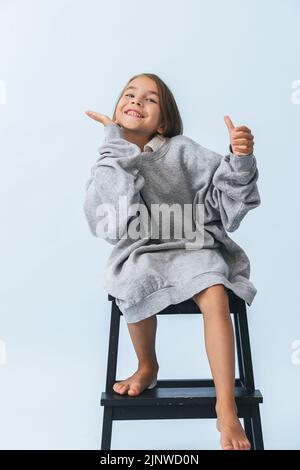 Une petite fille joyeuse et enjouée dans un gros sweat à capuche, assise sur un tabouret de marche Banque D'Images