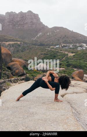 Vue arrière d'une femelle mince et flexible, debout sur un tapis et effectuant une fente en croissant avec révolution dans un paysage magnifique Banque D'Images