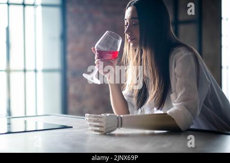 Une jeune fille calme et réfléchie tient un verre de vin dans le bras bionique à la maison, une femme avec un membre prothétique artificiel. Banque D'Images