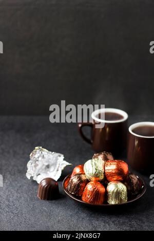 Bonbons au chocolat enveloppés dans du papier d'aluminium multicolore sur une assiette et deux tasses de café chaud sur fond noir avec espace de copie Banque D'Images