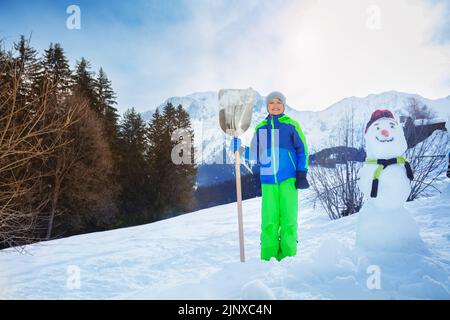 Stand de garçon avec pelle neige propre et la construction d'un bonhomme de neige Banque D'Images