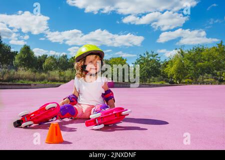 Petite fille adorable apprendre à skate rouleaux, s'asseoir dans le casque et sourire Banque D'Images