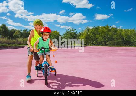 Deux enfants dans le garçon de parc enseignent à leur frère de faire du vélo Banque D'Images