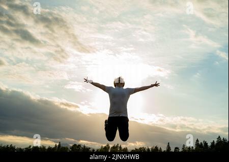 Vue d'en dessous d'un jeune homme excité sautant dans les airs avec ses bras larges ouverts sous ciel nuageux le soir. Banque D'Images