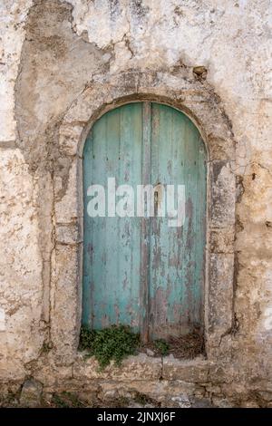 Porte pelée en bois vieilli fermée avec poignée en métal rouillé, façade de mur de bâtiment endommagée. Maison abandonnée, plante verte pousse sur l'escalier du ar bleu pâle Banque D'Images