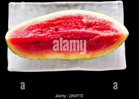 Une seule tranche de fruits frais et doux de pastèque placée sur un plateau blanc et isolée sur fond noir. Vue de dessus, gros plan. Banque D'Images