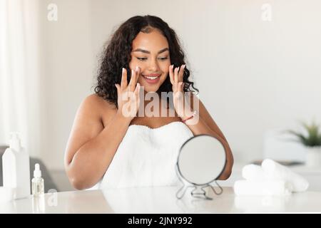 Concept de soin de la peau. Bonne femme noire surdimensionnée touchant son visage tout en regardant le miroir, appréciant la condition de peau Banque D'Images