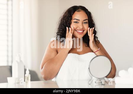 Belle femme noire plus grande taille dans une serviette faisant un massage de soulèvement du visage, assis à une table devant le miroir et souriant Banque D'Images