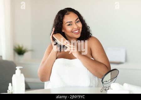 Bonne femme de chubby noir brossant ses cheveux bouclés avec le peigne et faisant la routine de soin des cheveux à la maison Banque D'Images