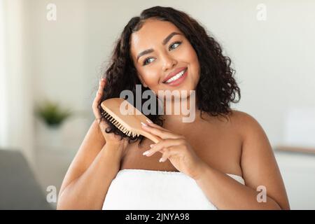 Soins capillaires domestiques. Bonne femme africaine américaine de corps se brossant les cheveux avec la brosse, debout dans l'intérieur de la salle de bains Banque D'Images