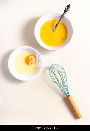 Œuf, coquille d'œuf avec fouet. Oeufs crus dans un verre et bol blanc sur la table. Pose à plat. Arrière-plan blanc cassé. Banque D'Images