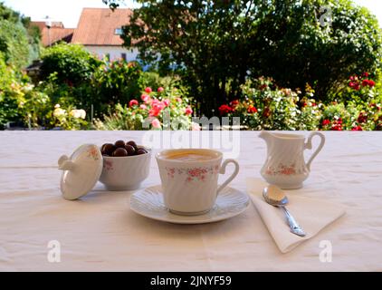 une belle tasse de café blanc chaud, un pot à lait et un bol de grains de chocolat sur la table recouverte d'une nappe blanche dans le jardin ensoleillé sur une somme Banque D'Images