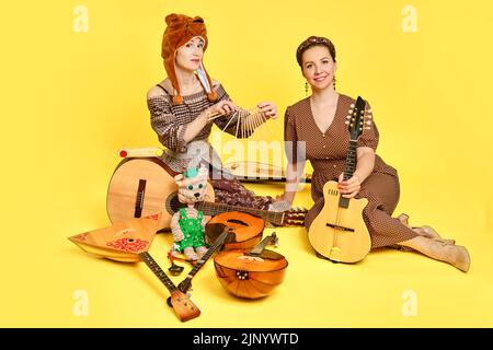 Femmes musiciens en robes avec instruments de musique sur fond de studio jaune. Des artistes heureux avec des instruments de musique à cordes avec des sourires sur le visage Banque D'Images
