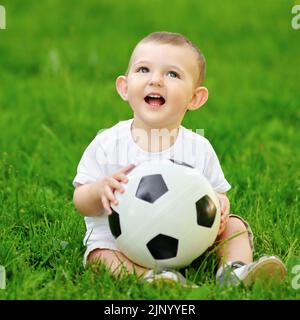 Un bébé garçon heureux joue avec un ballon de football sur l'herbe verte. Un enfant souriant en vêtements blancs est assis avec une balle dans ses mains, un an Banque D'Images