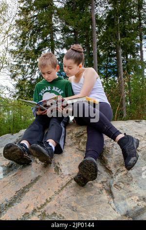 Issaquah, Washington, États-Unis. soeur de 11 ans et son frère de 9 ans lisant, la sœur l'aidant, assis sur un gros rocher dans un par Banque D'Images