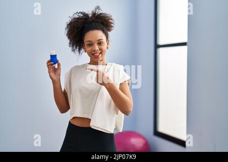 Jeune femme afro-américaine portant des vêtements de sport et une serviette tenant un déodorant souriant, souriant, pointant avec la main et le doigt Banque D'Images