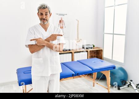 Thérapeute hispanique d'âge moyen, homme travaillant à la clinique de rétablissement de la douleur pointant des deux côtés avec les doigts, direction différente en désaccord Banque D'Images