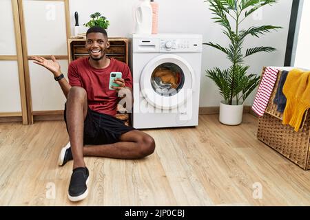 Jeune afro-américain utilisant un smartphone attendant le lave-linge pointant de côté avec les mains ouvertes palmiers montrant l'espace de copie, présentant advertisem Banque D'Images
